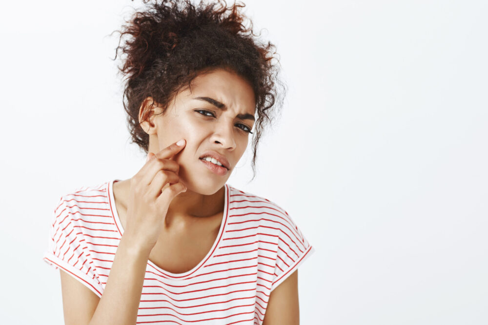 Displeased unhappy dark skinned woman with combed curly hair, frowning and pointing at acne on cheek, being upset with new pimple and bad face condition, standing disappointed over gray wall