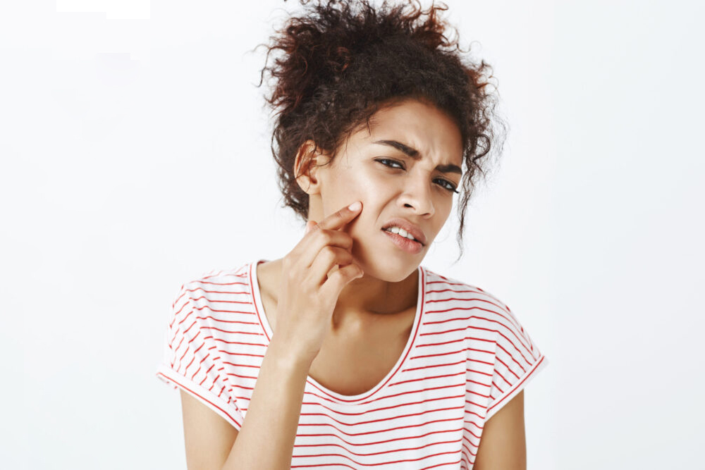 Displeased unhappy dark skinned woman with combed curly hair, frowning and pointing at acne on cheek, being upset with new pimple and bad face condition, standing disappointed over gray wall